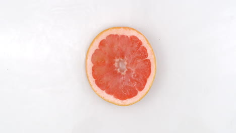 Top-view:-on-a-white-background-a-one-fresh-grapefruit-is-lying-in-the-water-water-drops-are-falling-from-above-and-splashes-are-falling-in-slow-motion-in-all-directions.